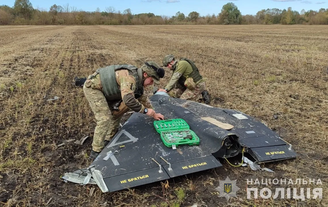 Двоє вибухотехніків поліції розбирають чорне трикутне крило збитого «Шахеда» на полі